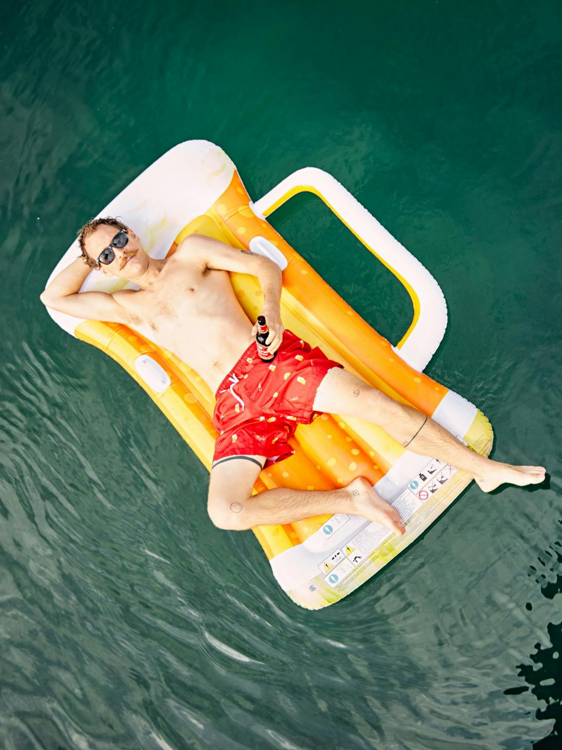 Junger Mann in Badehose von oben fotografiert liegt auf einer Luftmatratze in Form eines Bierglases © gm gobiq