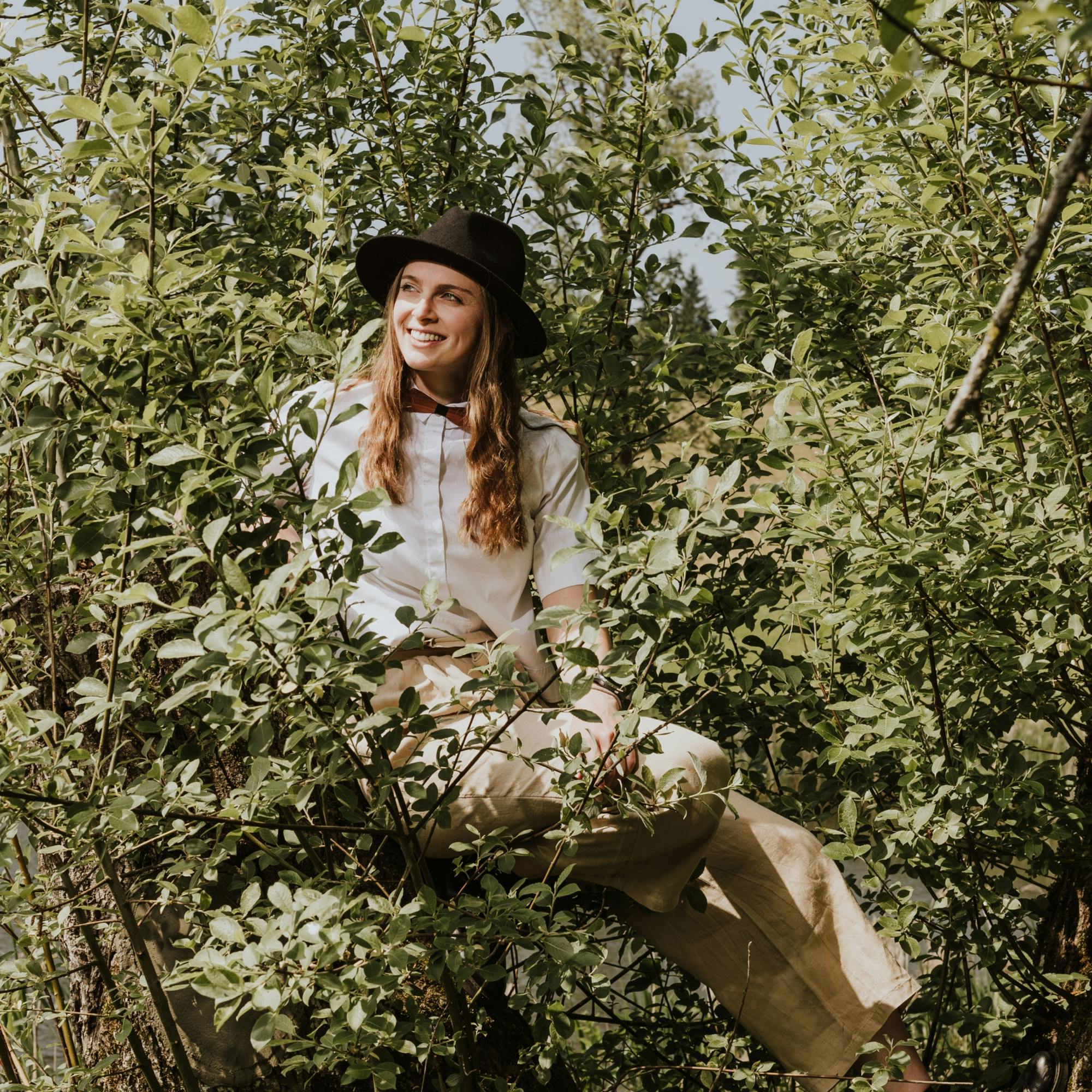 Junge Frau im Bohemian Look trägt eine Holzfliege und einen Hut und klettert im Baum © good matters