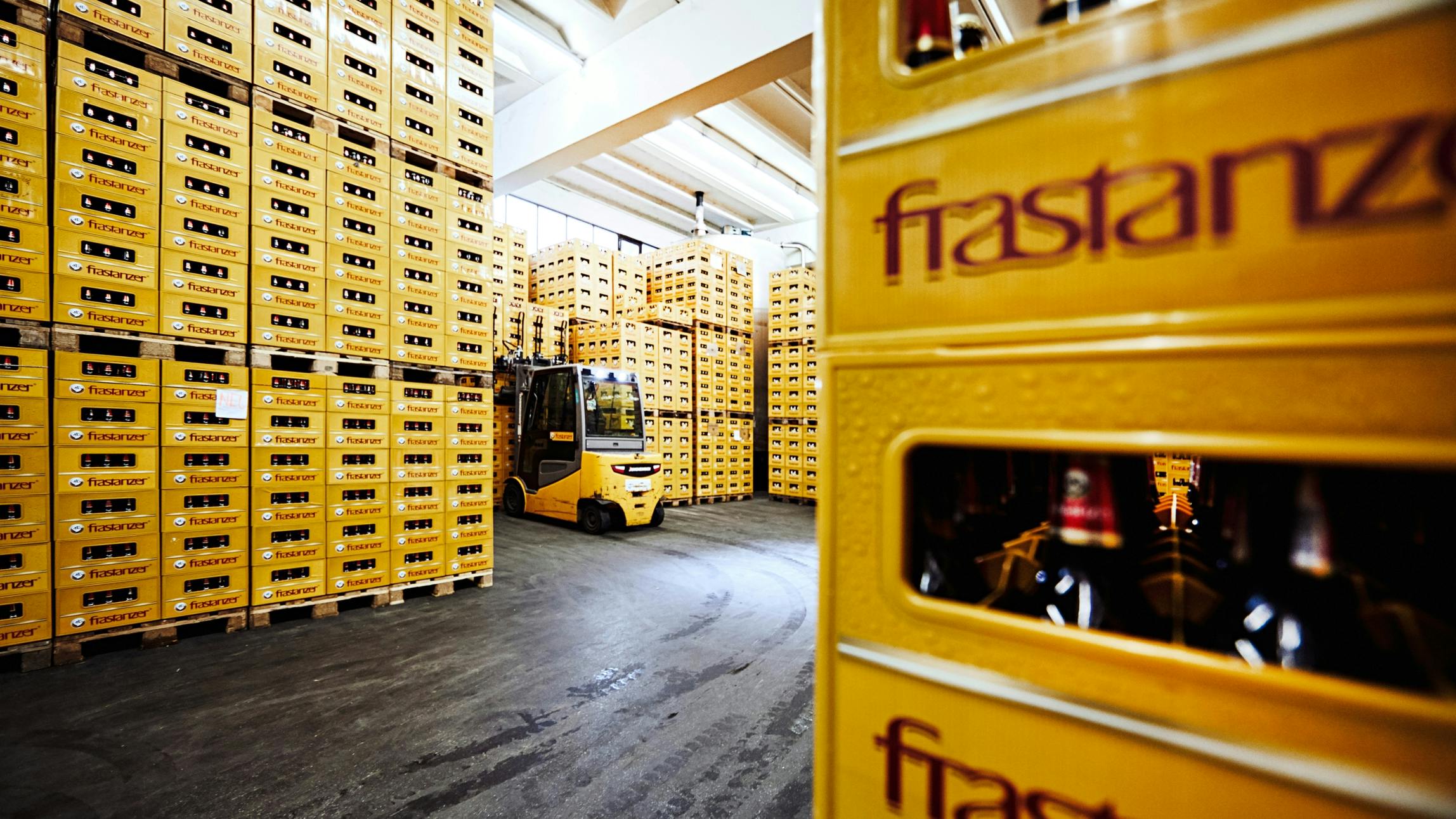 Die Lagerhalle der Frastanzer Brauerei mit Paletten voll gelben Bierkisten. Ein Staplerfahrer hebt gerade eine Palette vom Stapel. © gobiq