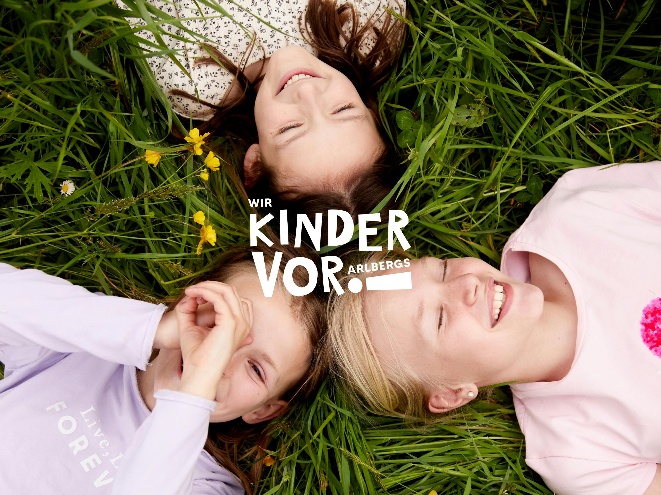 Drei Mädchen liegen in einer Wiese, eine hält die Hände vor das Gesicht wie ein Kaleidoskop. Das Wir Kinder Vorarlbergs Logo im Vordergrund. © gobiq