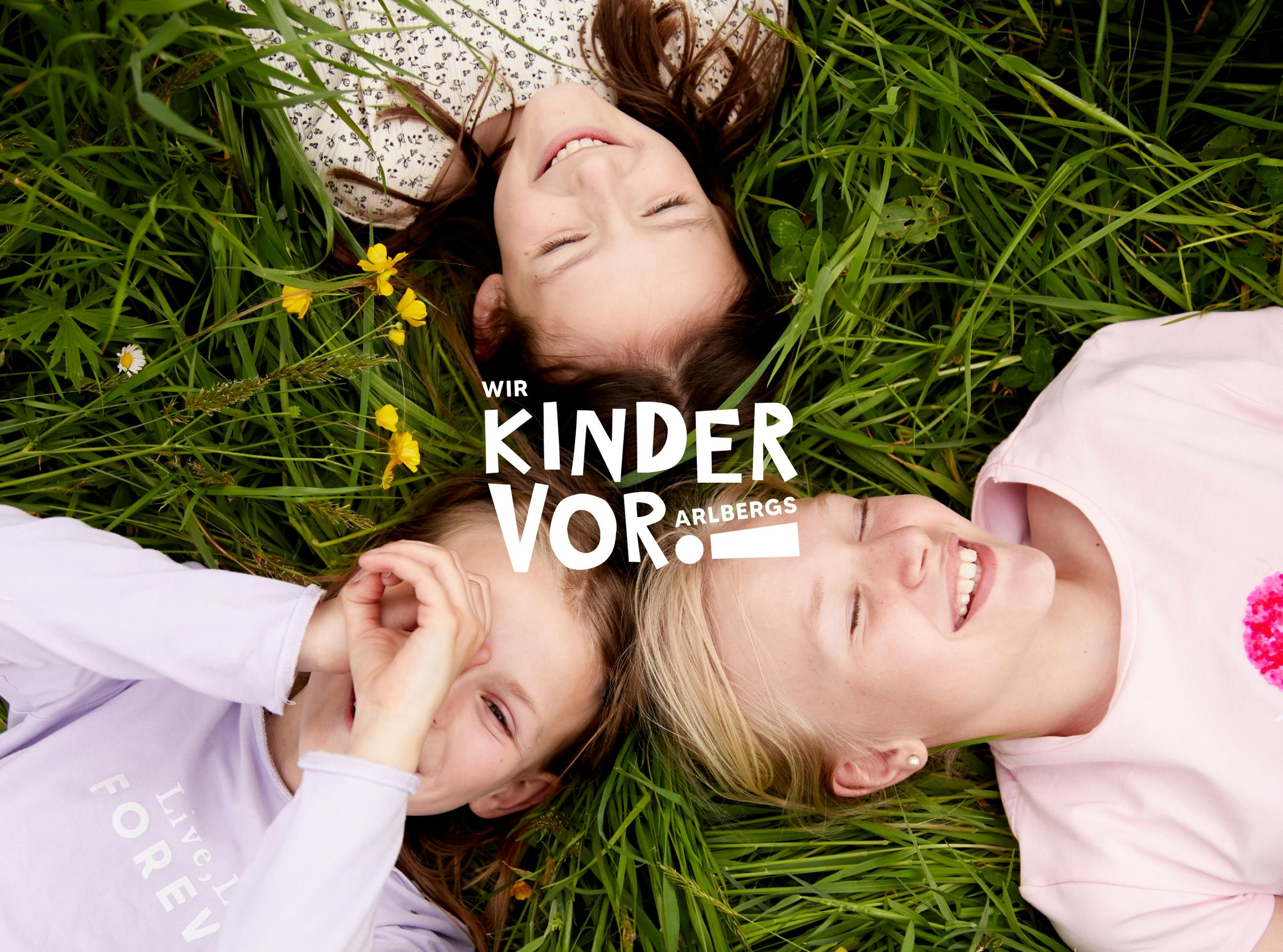 Drei Mädchen liegen in einer Wiese, eine hält die Hände vor das Gesicht wie ein Kaleidoskop. Das Wir Kinder Vorarlbergs Logo im Vordergrund. © gobiq