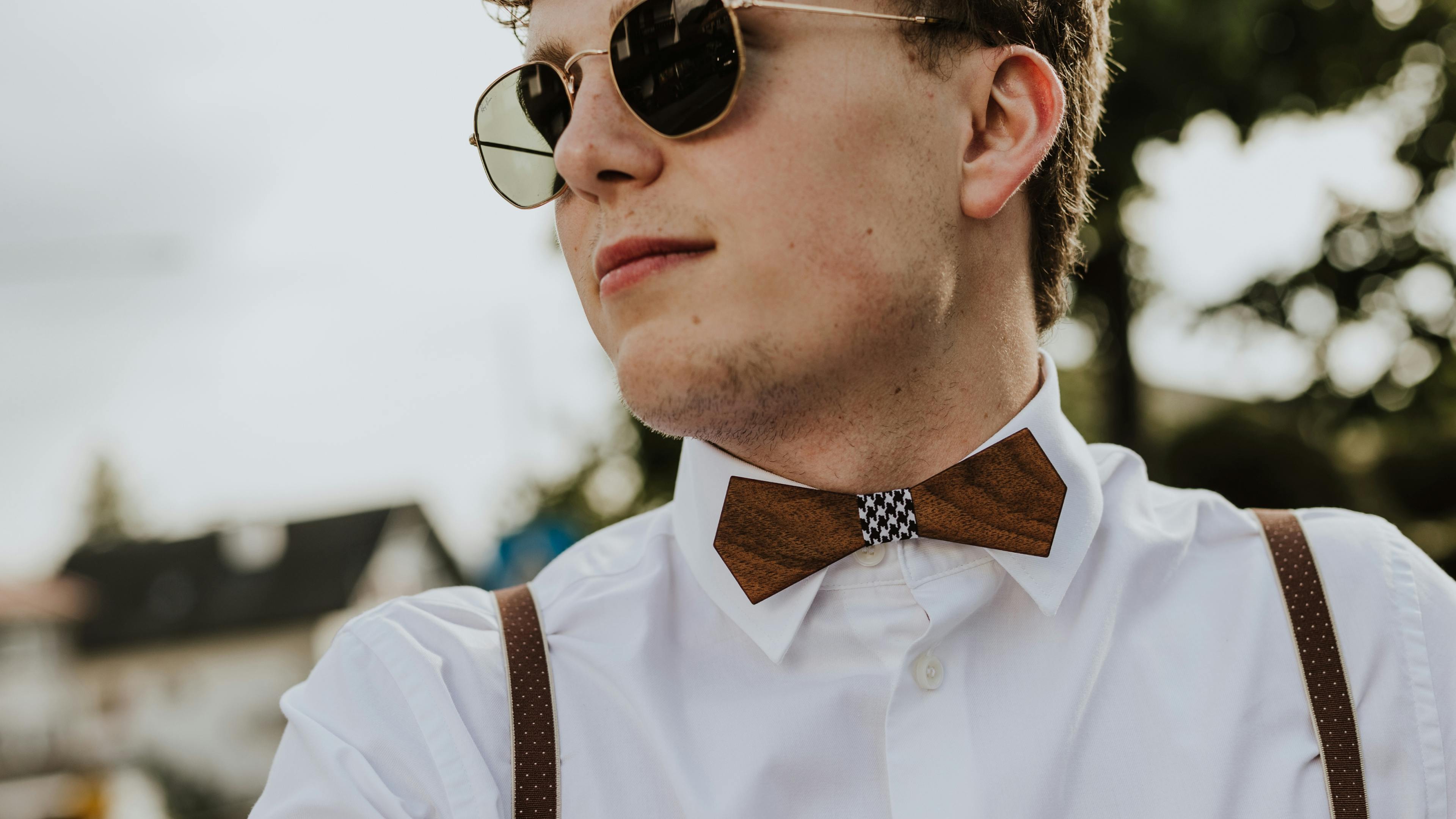 Junger Mann mit cooler Sonnenbrille und Hosenträgern trägt Holzfliege © good matters