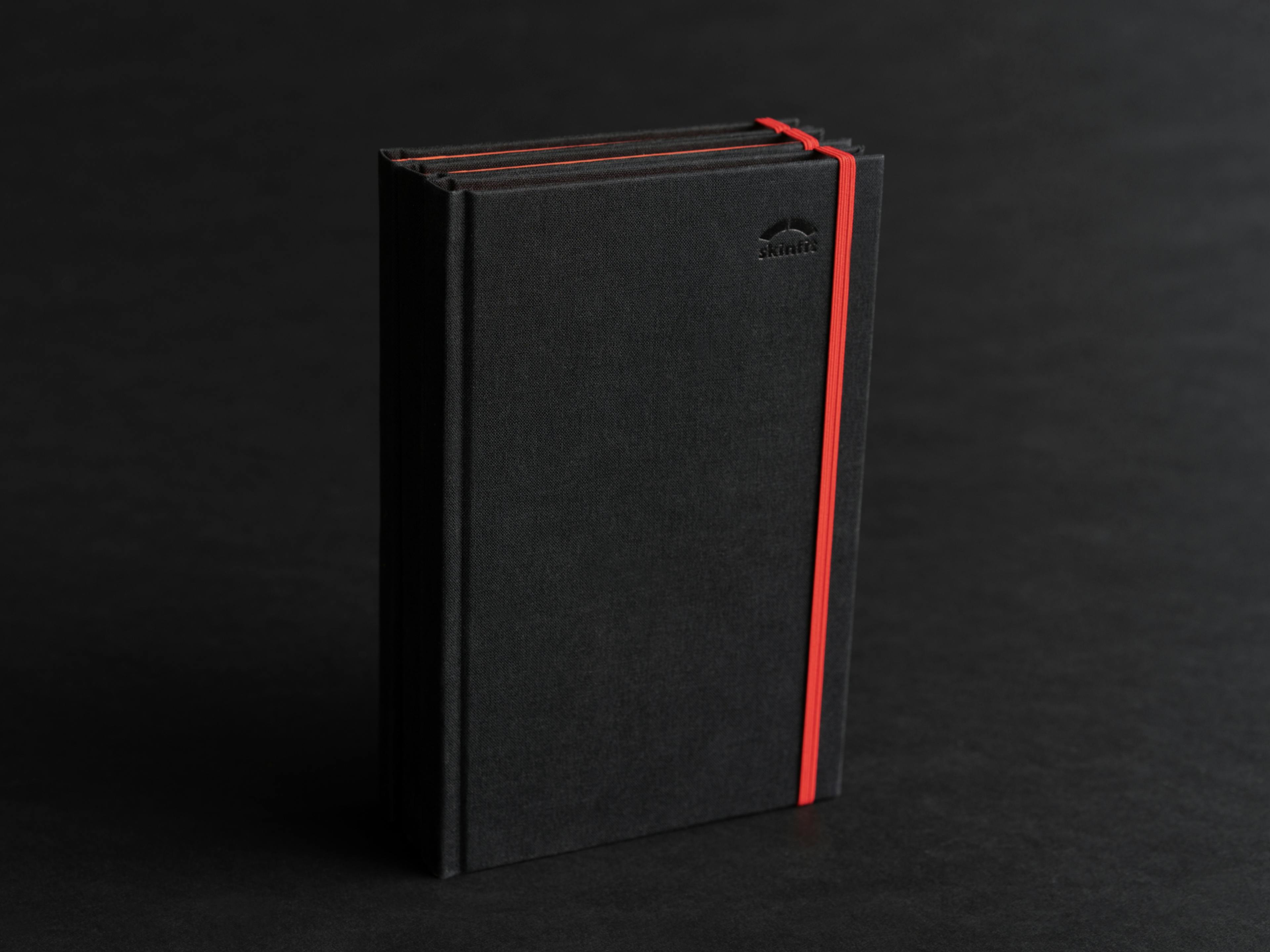 Foto von 3 skinfit brandbooks auf schwarzem Hintergrund, schwarzer Leineneinband, Blindprägung und rotes Gummiband © gm gobiq