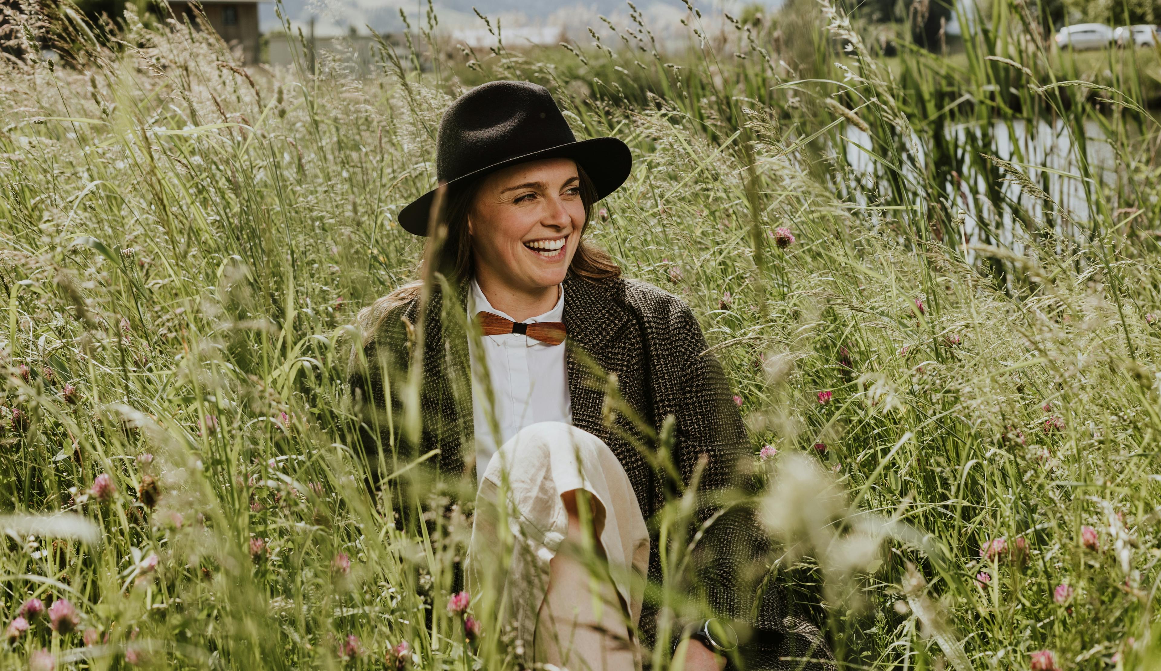 Junge Frau im Bohemian Look trägt Hut und eine schicke Holzfliege und sitzt lachend in einer wilden Wiese © good matters